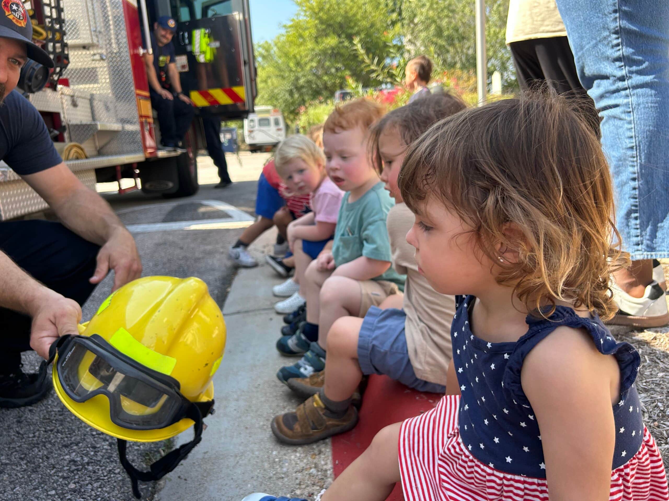 Preschool children sitting on a curb looking at a firefighter’s yellow helmet and goggles beside a fire truck during Fire Safety Week at Primrose School of Four Points in Austin, Texas