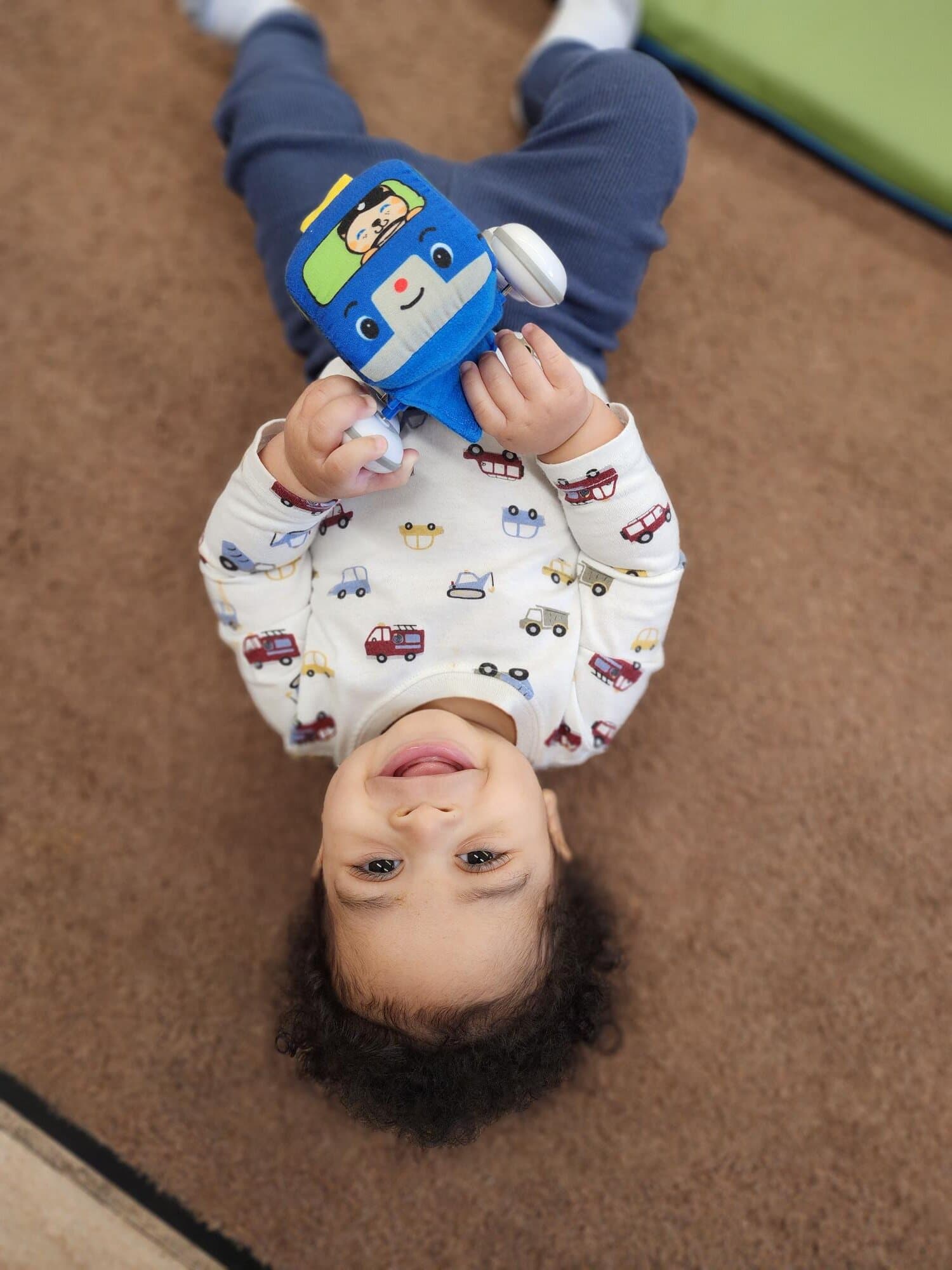 Smiling baby lying on a carpet and holding a blue toy vehicle, looking up at the camera.