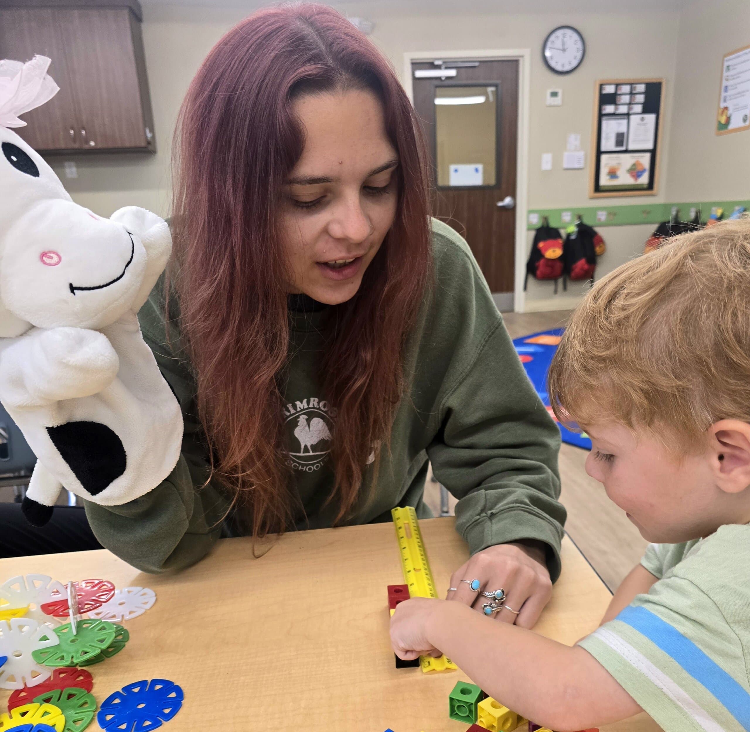 Toddler measuring blocks with a teacher using a tape measure during a hands-on learning activity at a daycare in Keller, Texas, with a cow puppet used for playful engagement.