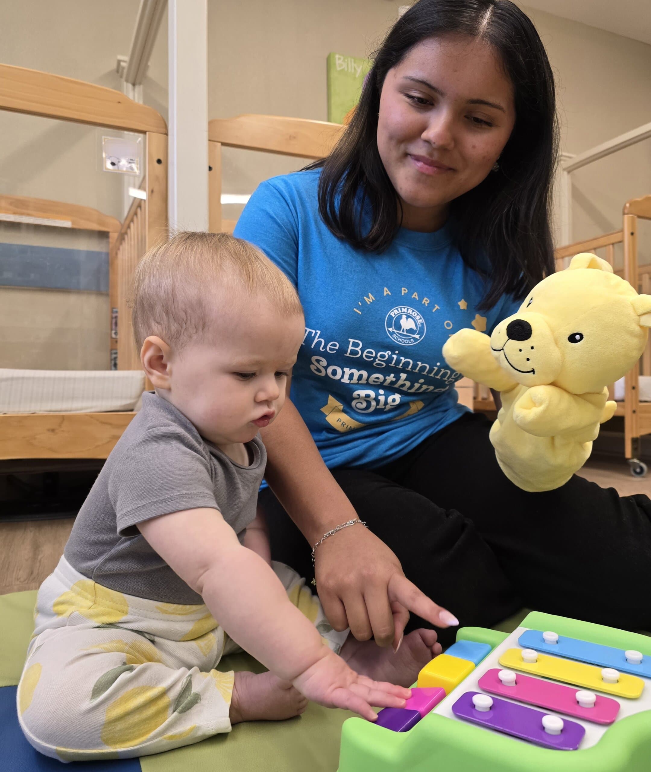 Infant playing a colorful toy piano with a teacher and Erwin puppet at a daycare in Keller, Texas, supporting early development through music and sensory play.