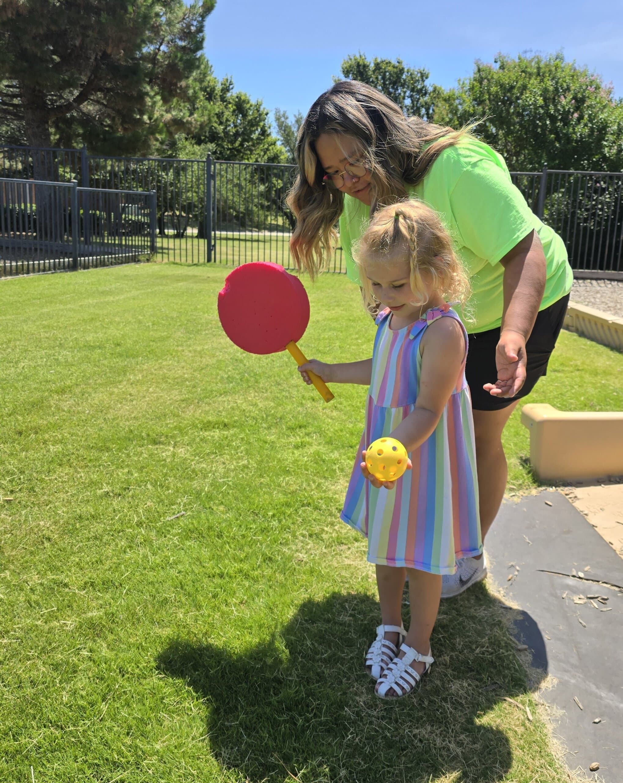 Toddler girl playing paddle ball with a teacher on the playground at a daycare in Keller, Texas, enjoying outdoor learning and motor skill activities.