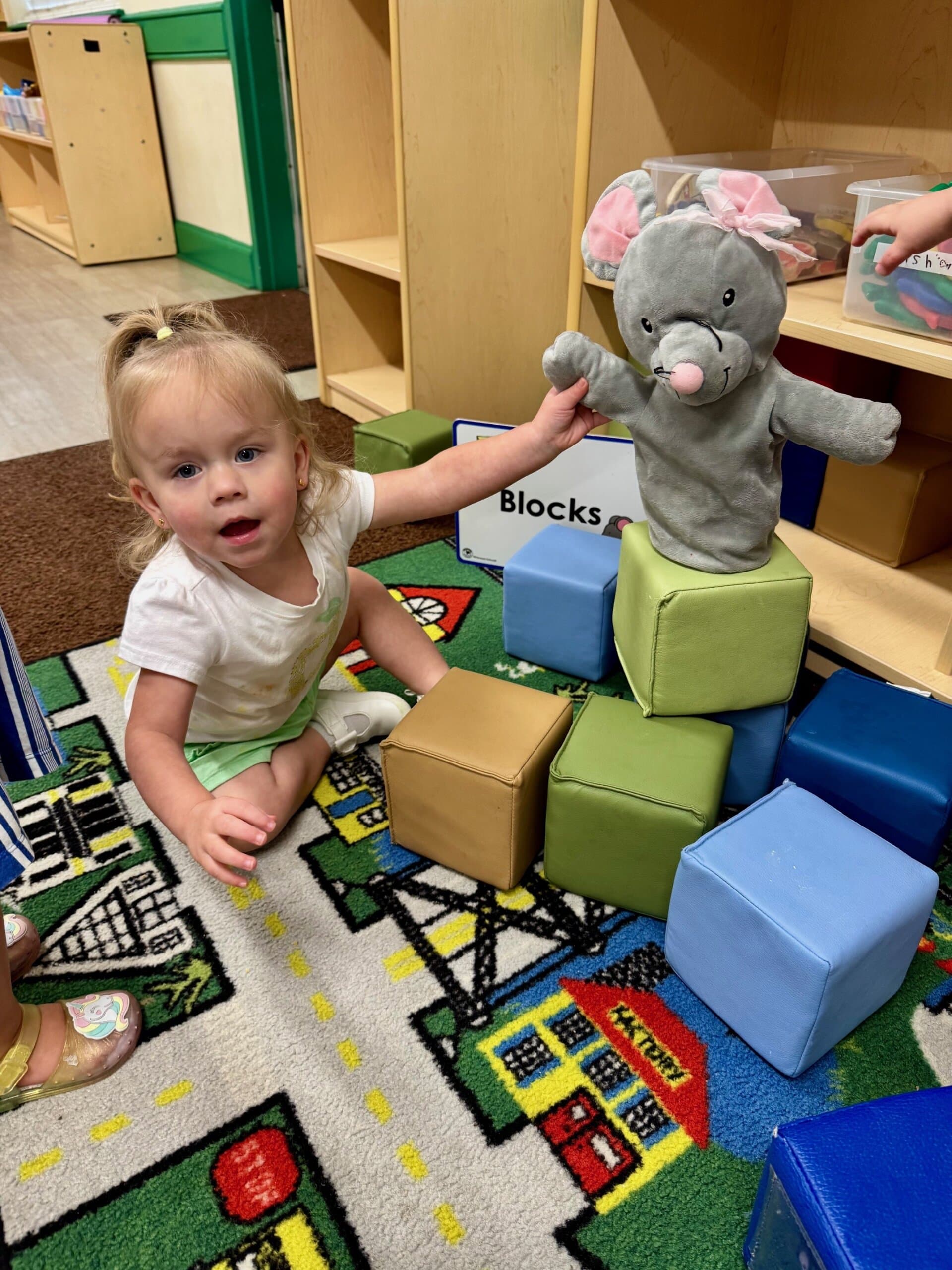 Toddler building in the block center while holding a puppet, engaging in imaginative and constructive play that supports creativity and motor skills