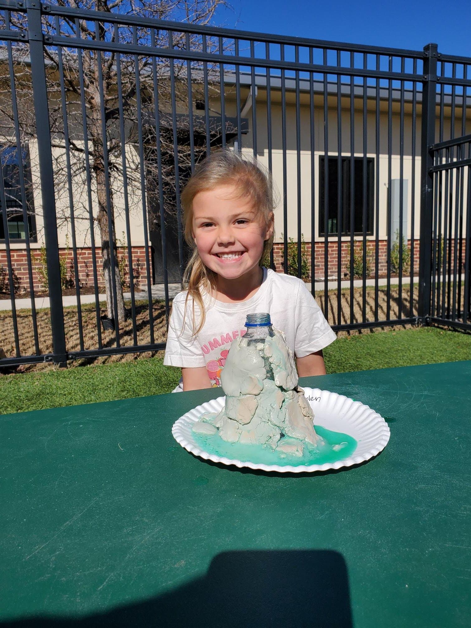 Smiling preschool girl at Primrose School sitting outdoors with a homemade volcano science experiment made of clay and a water bottle, showcasing STEM learning and hands-on discovery