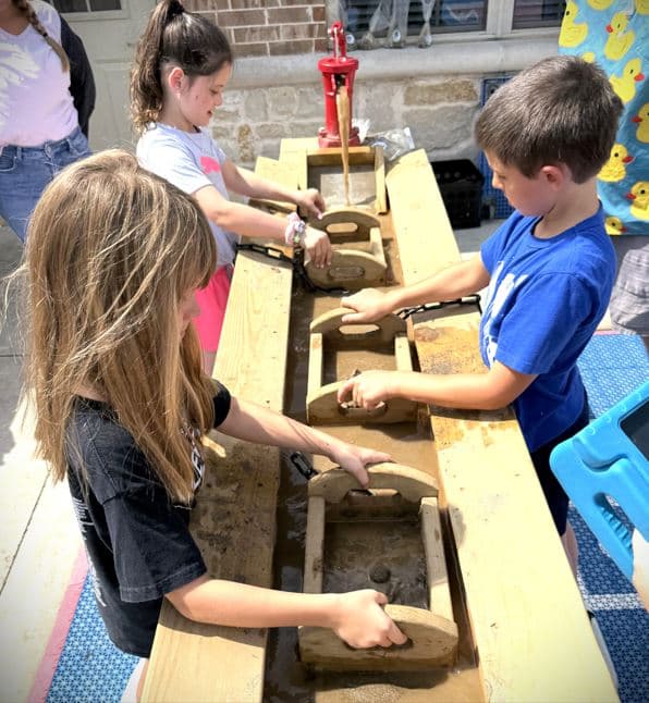 School aged students using wooden sifters in water the uncover gems for an in-house field trip during summer camp