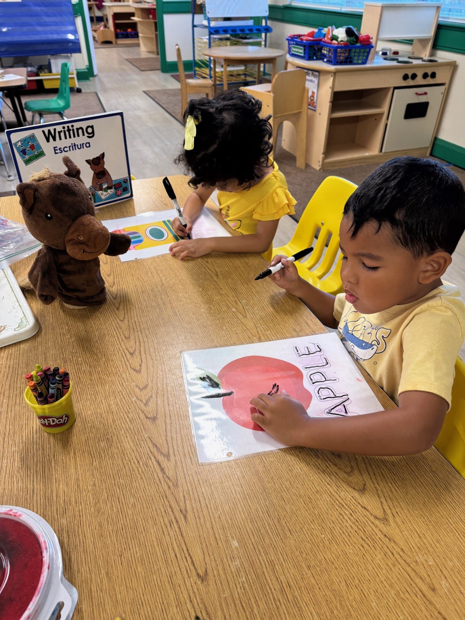 Children at a writing center tracing letters with dry erase markers and worksheets, developing early writing skills