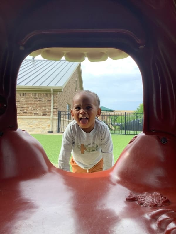 Toddler smiling and crawling through a playground tunnel at Primrose School at Alliance in Fort Worth, Texas. Outdoor play supporting physical development, exploration, and joyful learning in a safe preschool environment.