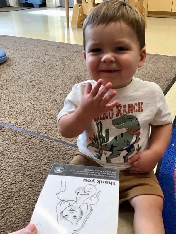 Toddler boy signing thank you with a sign language card of thank you in front of him