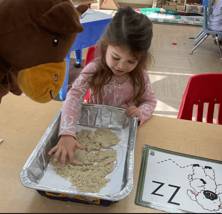Preschool student tracing the letter Z in sensory sand while participating in a hands-on alphabet learning activity with a classroom character puppet.