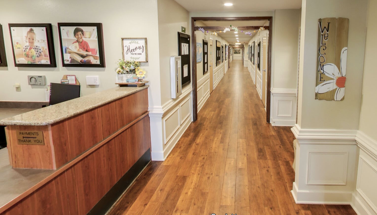 The welcoming reception area and hallway at Primrose School of Eagan, featuring hardwood floors, children's artwork displays, and classroom doors extending down a bright, well-appointed corridor.
