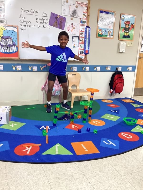 Student proudly standing beside a colorful block structure they designed and built during a hands-on STEM and engineering activity in the classroom.