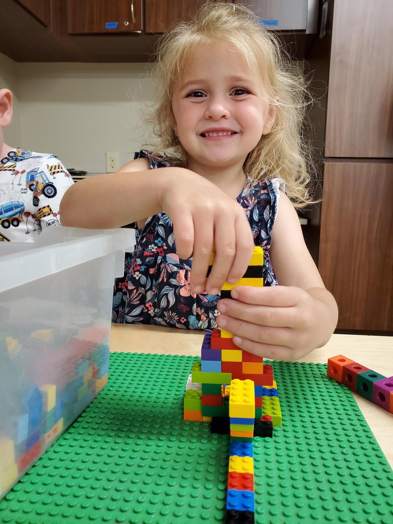 Smiling preschool girl at Primrose School building with colorful interlocking blocks on a green baseplate, engaging in hands-on STEM learning and creative play.