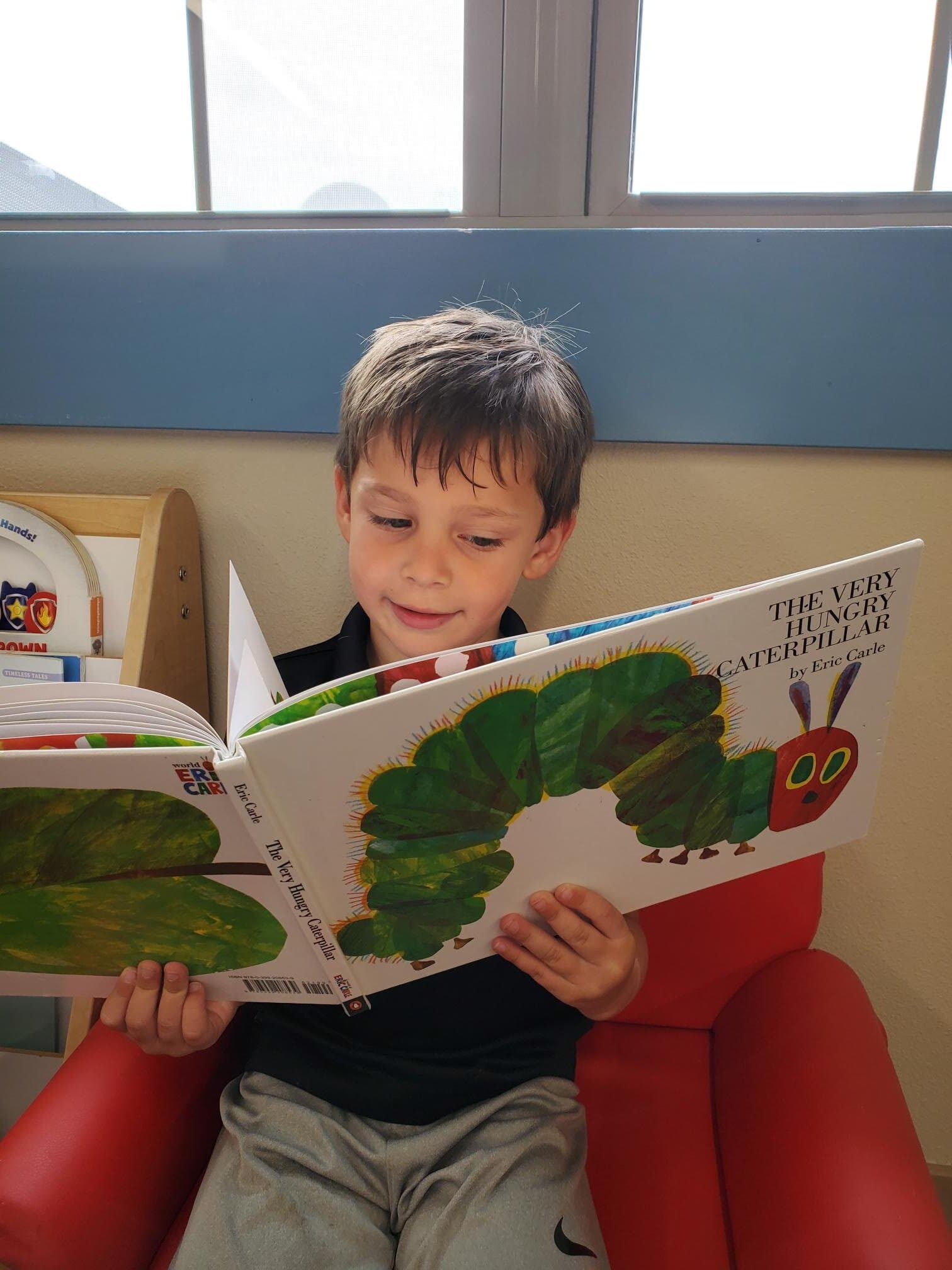 Preschool boy at Primrose School at Alliance reading The Very Hungry Caterpillar by Eric Carle while sitting in a cozy reading corner, promoting early literacy and a love of books.