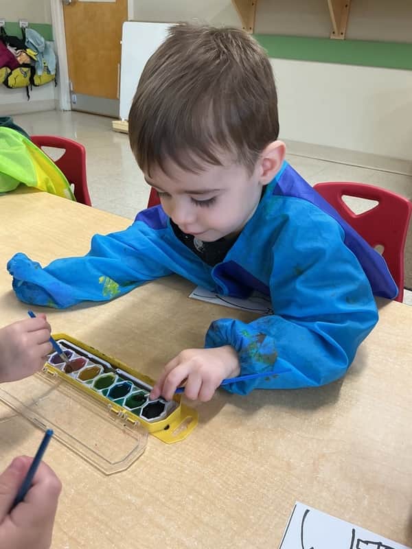 A boy in a smock painting with watercolors at a table.