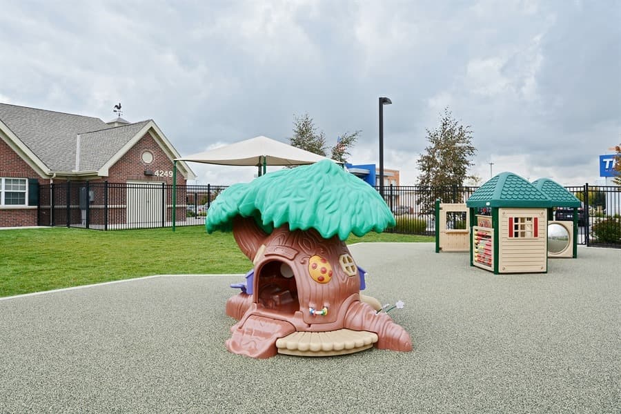 Early Preschool outdoor play area at Primrose School of Eagan featuring a whimsical tree house play structure, interactive activity panels, rubber safety surfacing, and secure black iron fencing