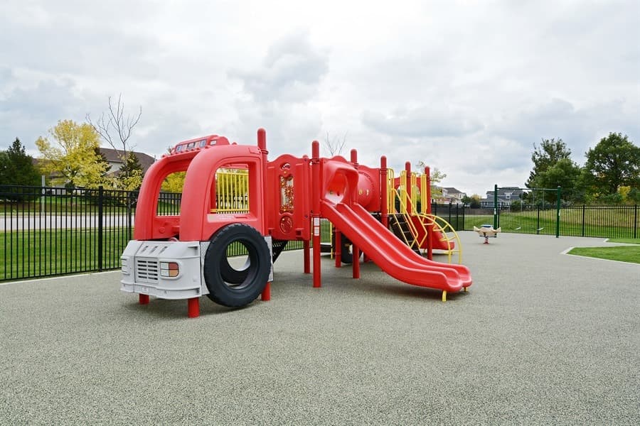 Preschool Pathways outdoor play area at Primrose School of Eagan featuring a large red fire truck play structure with dual slides, climbing elements, and rubber safety surfacing within a secure black iron fence