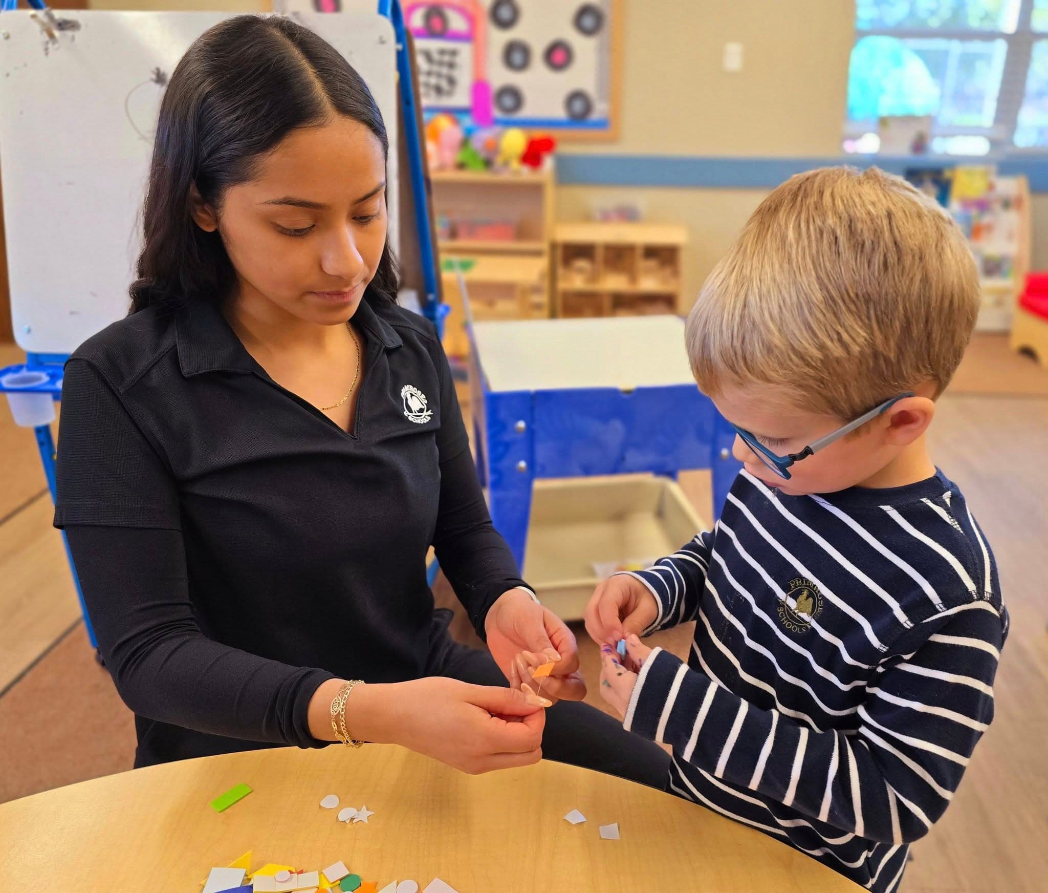 Primrose School teacher in Fort Worth helping a preschool boy with a hands-on fine motor skills activity using small building blocks at a classroom table, supporting early childhood development and learning through one-on-one guidance in Heritage, Texas