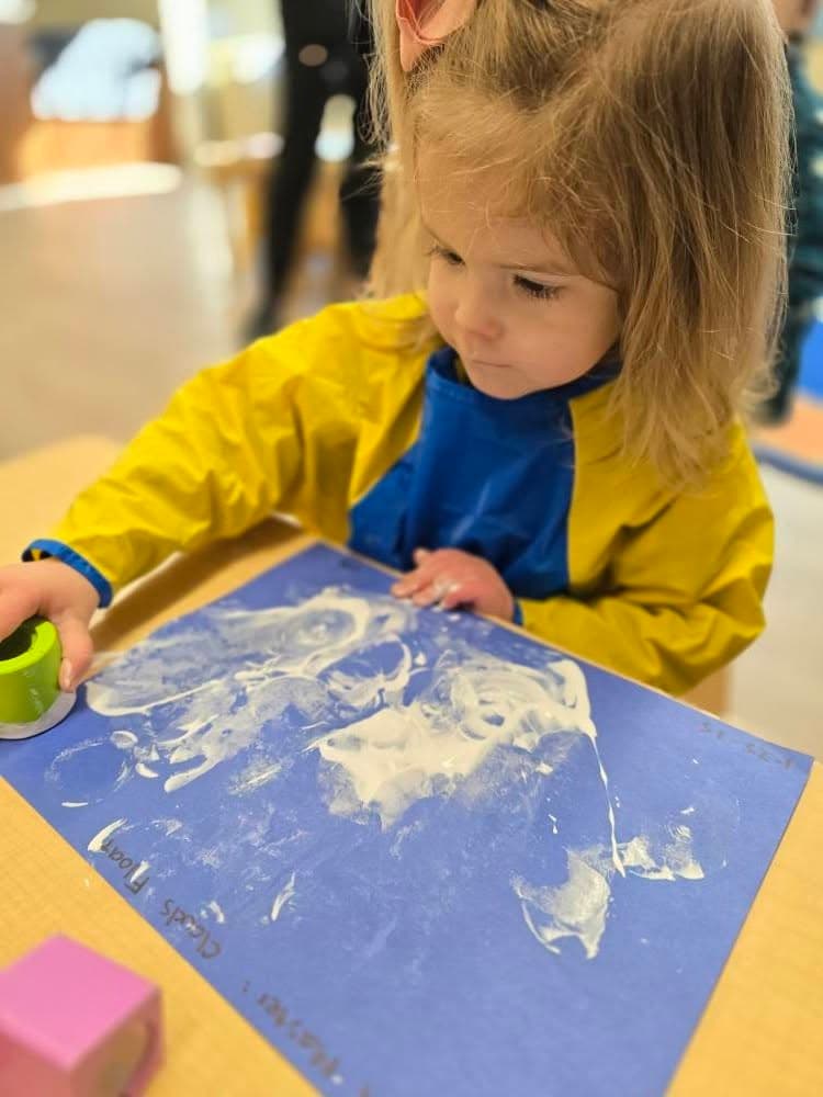 Preschool-aged child in a yellow smock engaged in a sensory painting activity with white paint on blue paper at a table.