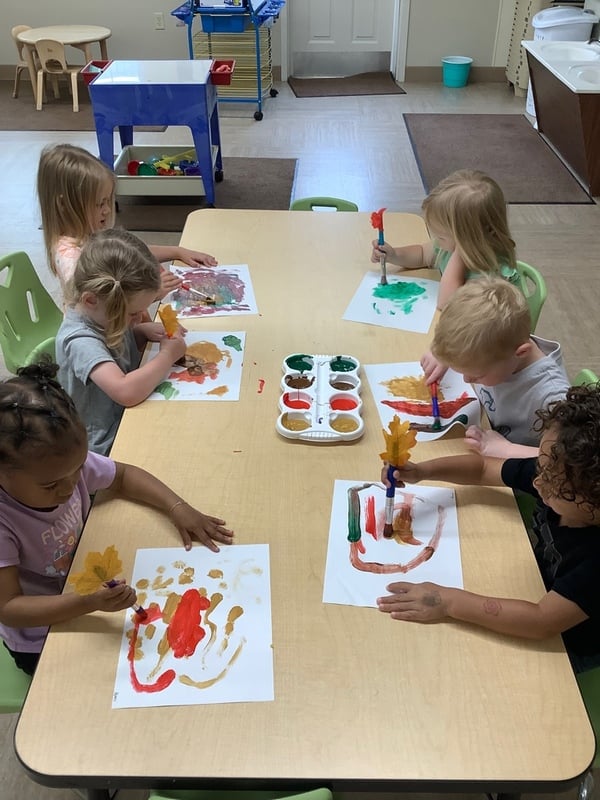 Preschool children at Primrose School painting with brushes at a classroom table, engaging in creative expression and fine motor development during a group art activity.
