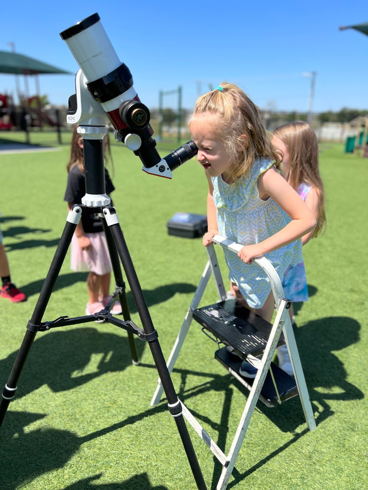 Young girl climbing a step ladder to look through a telescope during outdoor school STEM activity at Primrose NW Fort Worth