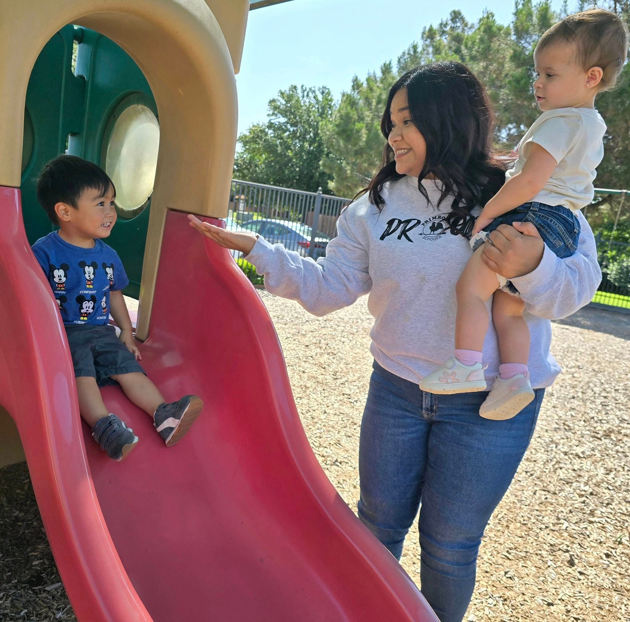 Caregiver helps preschooler and toddler on playground slide during outdoor play to develop social and gross motor skills.