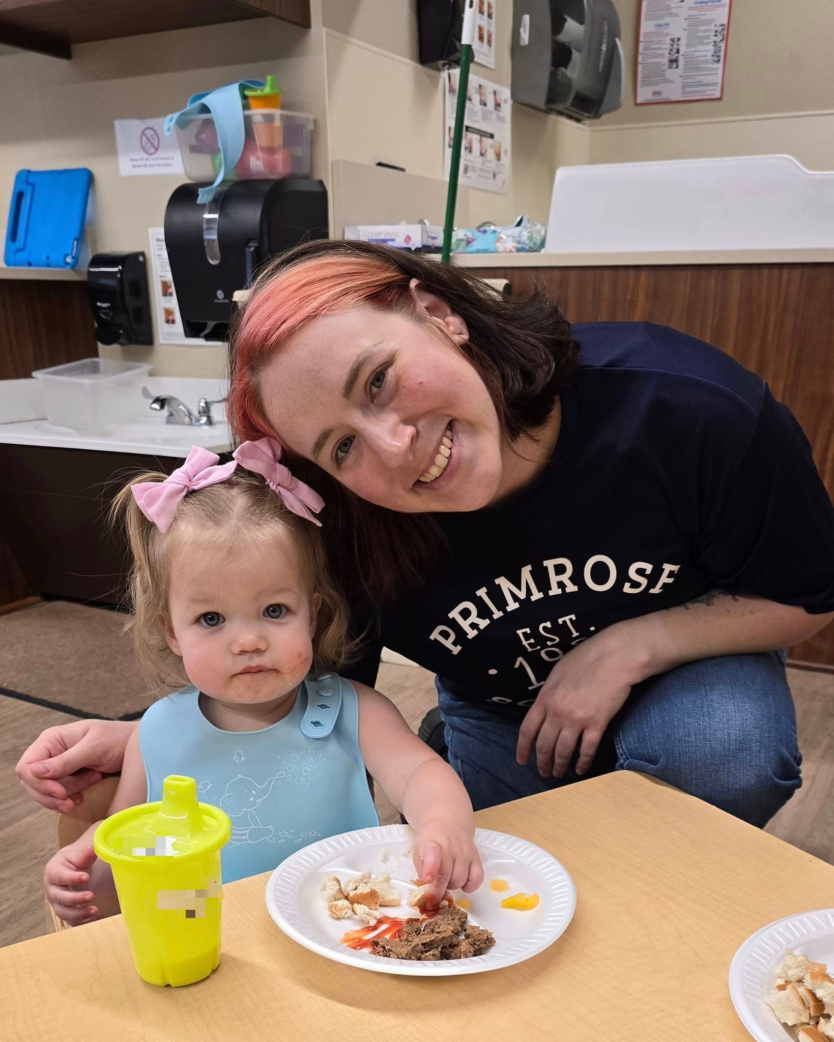 Preschool teacher smiling next to a young toddler enjoying a meal at the table during a classroom mealtime, promoting healthy eating habits and nurturing connections at the best childcare facility in Heritage.