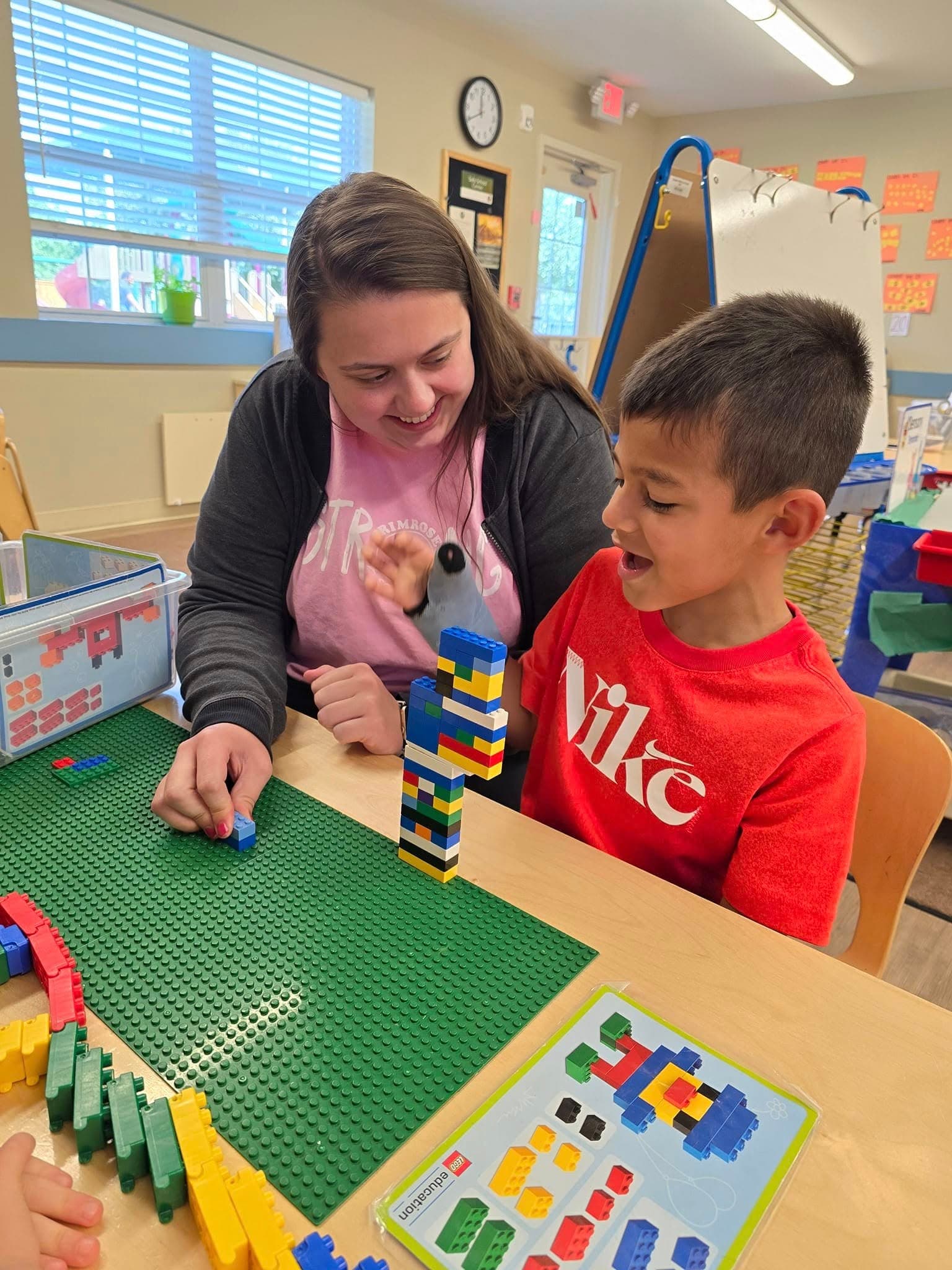 Teacher and PreK child building LEGO tower at table to develop fine motor and early STEM skills in classroom