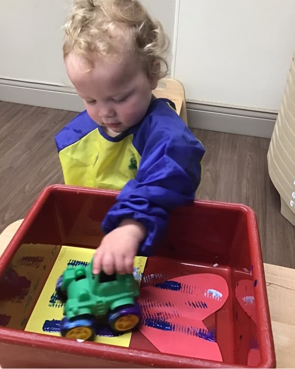 Toddler engaging in a sensory art activity at Primrose School of NW Fort Worth in Haslet, TX, using a toy truck to create paint tracks in a discovery bin. Creative expression, fine motor development, and hands-on learning in a nurturing preschool environment.