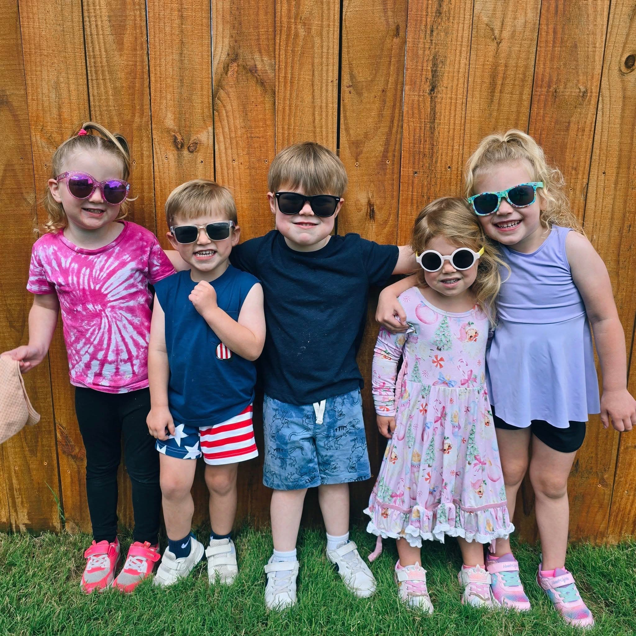 Group of preschool children wearing sunglasses and summer outfits, smiling and standing in front of a wooden fence at a childcare center in Keller, Texas.