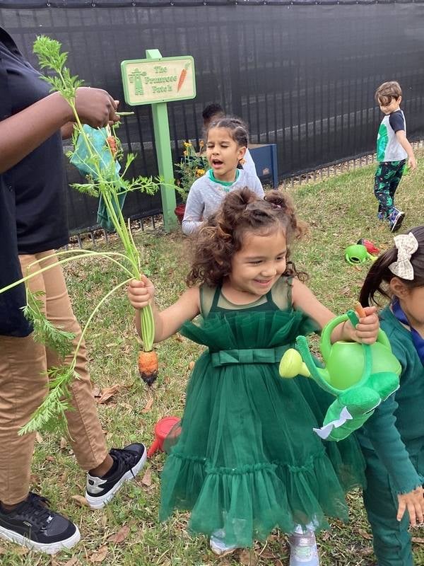 child in green dress outside holding carrots from the garden and a watering can
