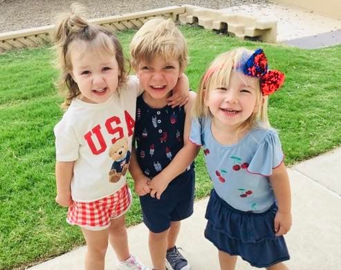 three early preschool children posing together in red white and blue patriotic fourth of july outfits on the playground