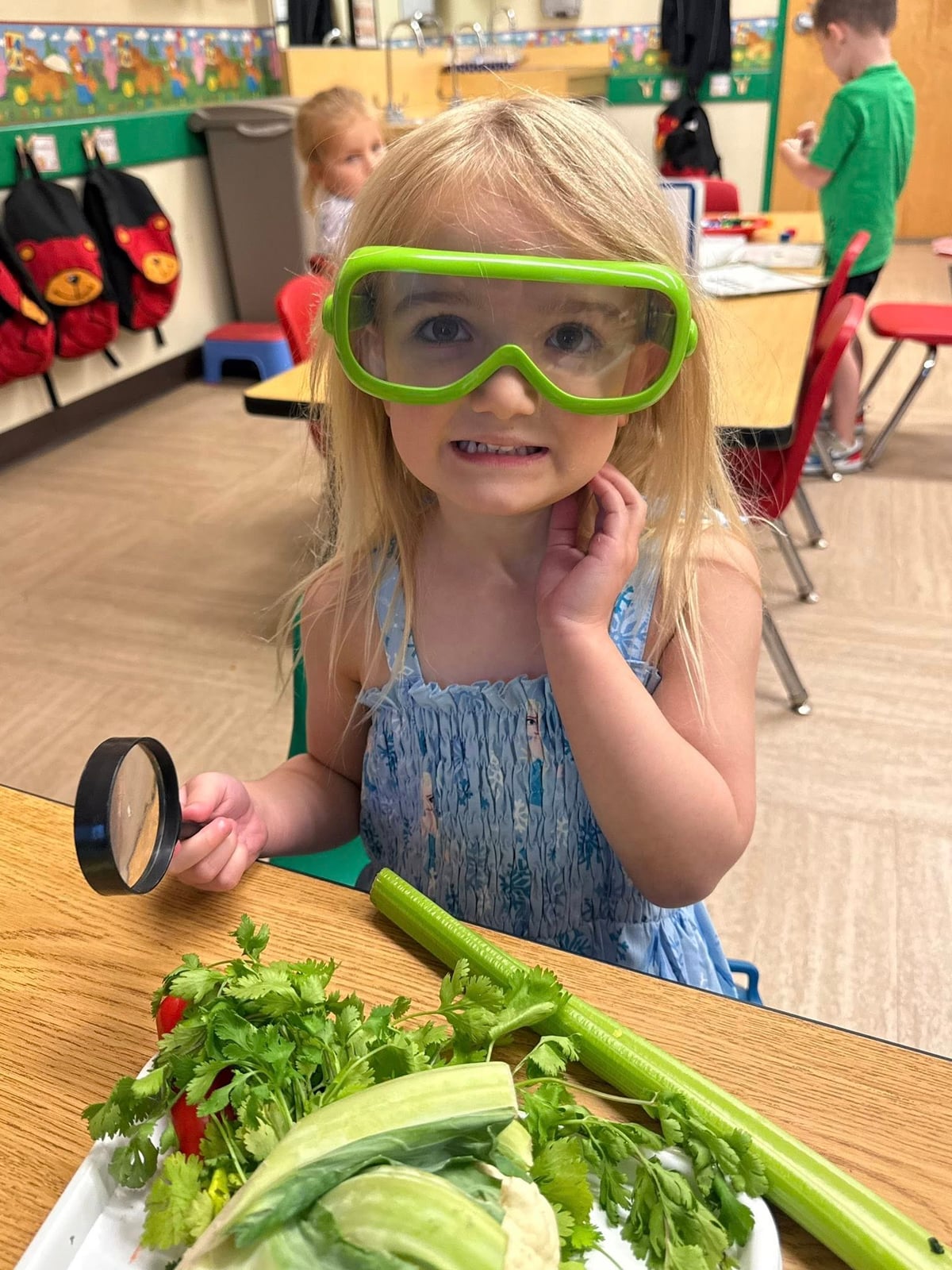 Child wearing safety goggles and using a magnifying glass to investigate vegetables during a hands-on science and discovery lesson.