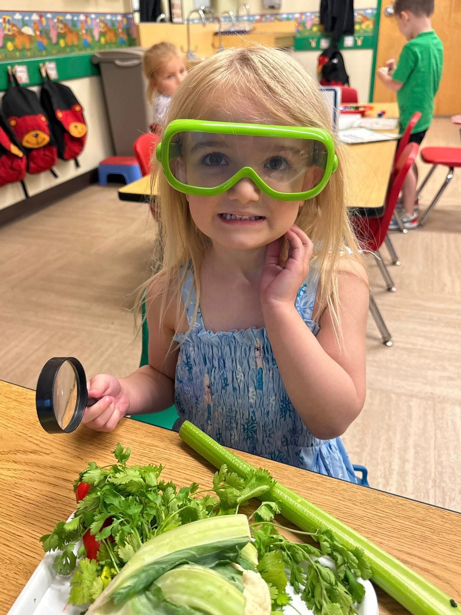 Child wearing safety goggles and using a magnifying glass to investigate vegetables during a hands-on science and discovery lesson.