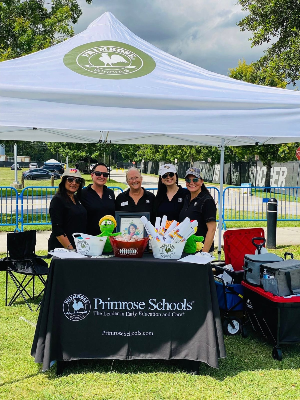 White tent with green primrose logo 5 women standing in green grass behind a black tablecloth