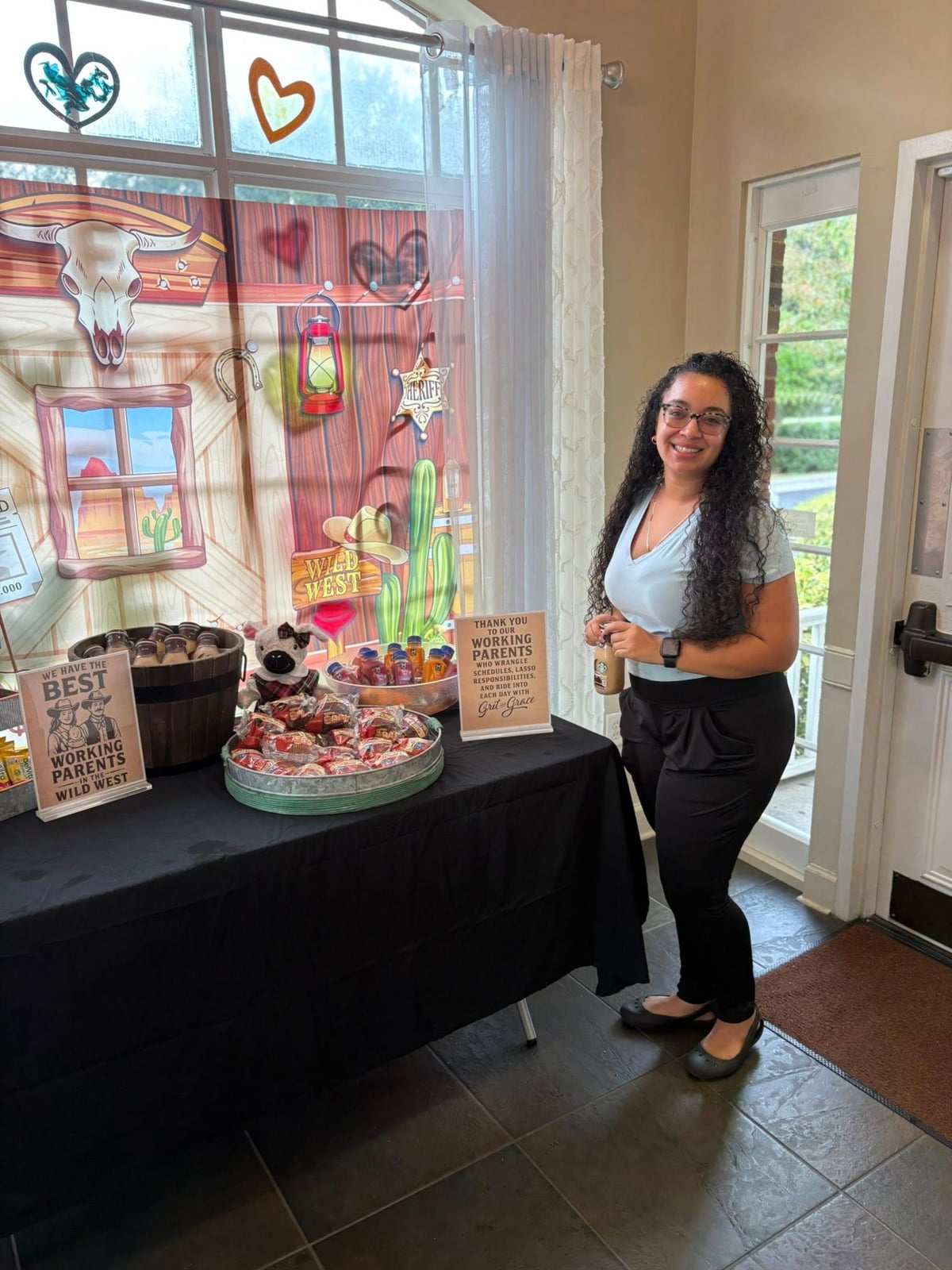 Woman in black pants and blue top with long dark hair holding a coffee in front of a table with a black tablecloth with food on top of it to grab and go, Pink western backdrop behind the table