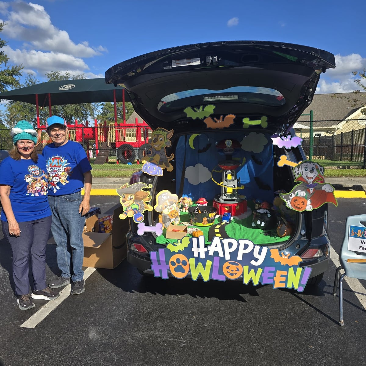 Trunk decorated with dogs that says happy howlloween and two adults in blue shirts standing next to it