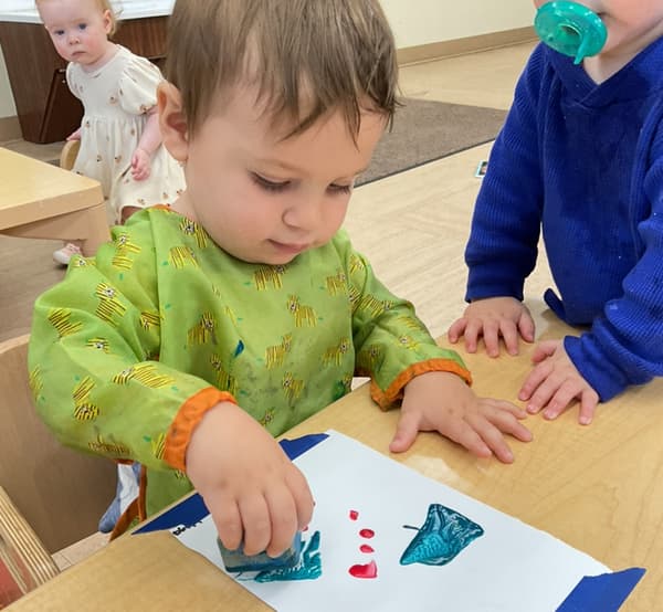 Toddler creating artwork with paint and stamping materials while developing fine motor skills, creativity, and self-expression during a classroom activity.