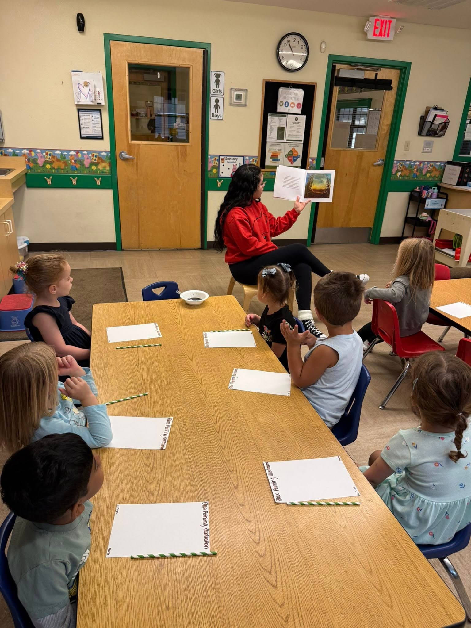 Preschool students gathered around a teacher during story time, listening attentively as a book is read aloud.