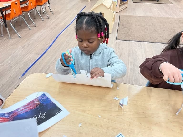 Young child carefully cutting paper with safety scissors during a classroom activity that develops hand strength, coordination, and early fine motor skills.