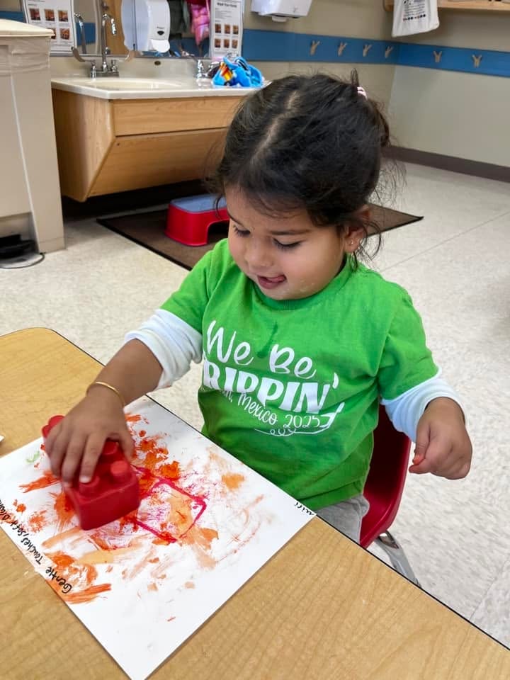 Toddler using a large block to stamp colorful paint onto paper during a process art activity that builds creativity, sensory exploration, and fine motor skills at Primrose School.