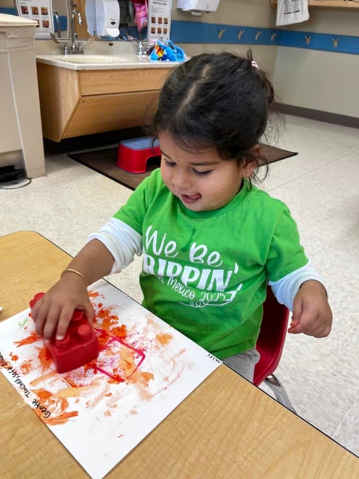 Toddler using a large block to stamp colorful paint onto paper during a process art activity that builds creativity, sensory exploration, and fine motor skills at Primrose School.