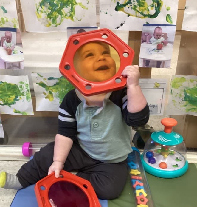 Infant holding a red framed mirror during floor play at Primrose School of Thornton