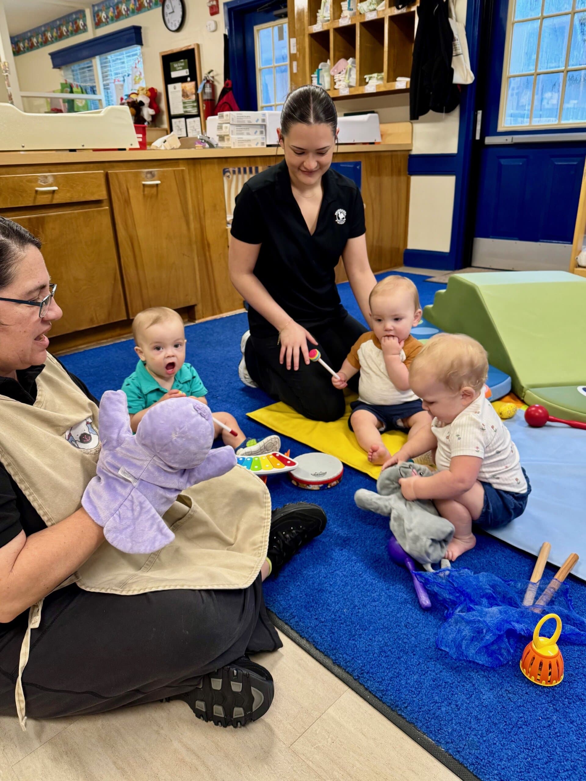 Infants playing musical instruments with guidance from teachers in a classroom setting, promoting early childhood development and music education.