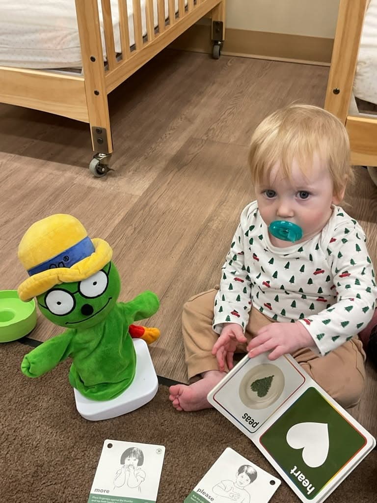 Infant sitting on the classroom floor holding communication cards and learning materials while interacting with Og® the Bookworm during a language and social-emotional learning activity.