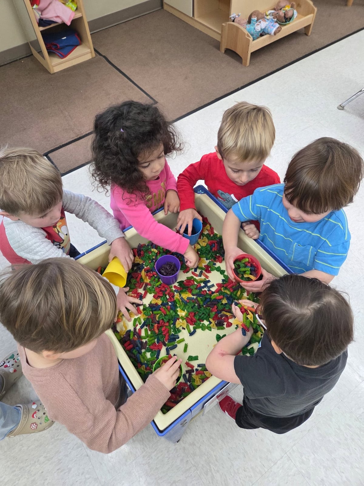 Preschoolers using cups and scoops to explore a colorful pasta sensory bin during a hands-on classroom activity that promotes sensory exploration, cooperation, counting, and fine motor development at Primrose School.
