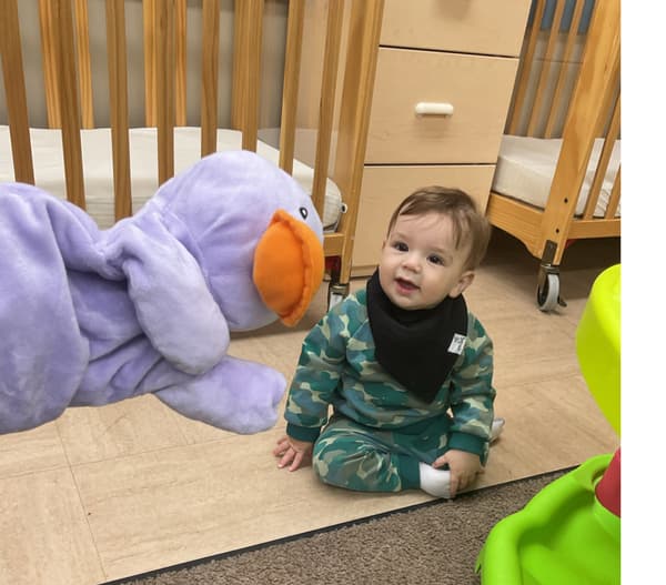 Infant sitting on the classroom floor interacting with a purple plush puppet during a social-emotional learning activity.