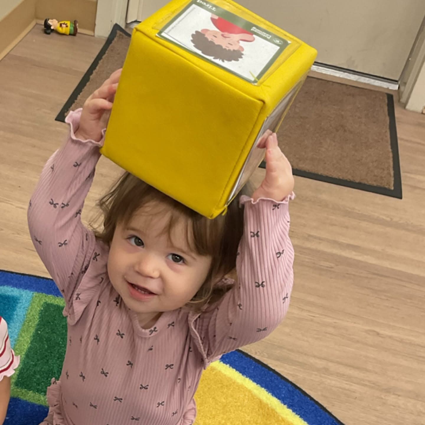 Toddler balancing a learning cube on her head while exploring spatial awareness, problem-solving, and imaginative play in the classroom.