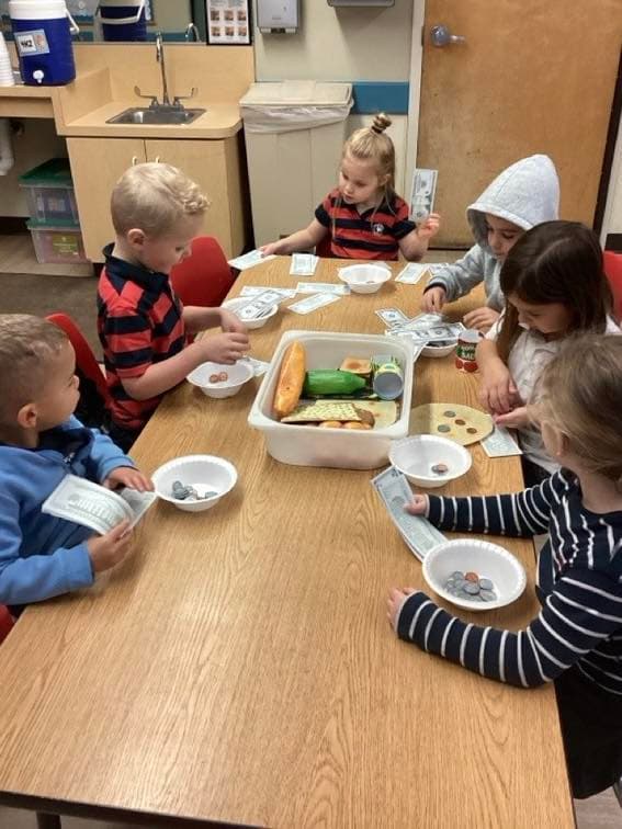 Preschool students participating in a classroom grocery store activity, using play money and food items to practice counting, decision-making, and social skills.