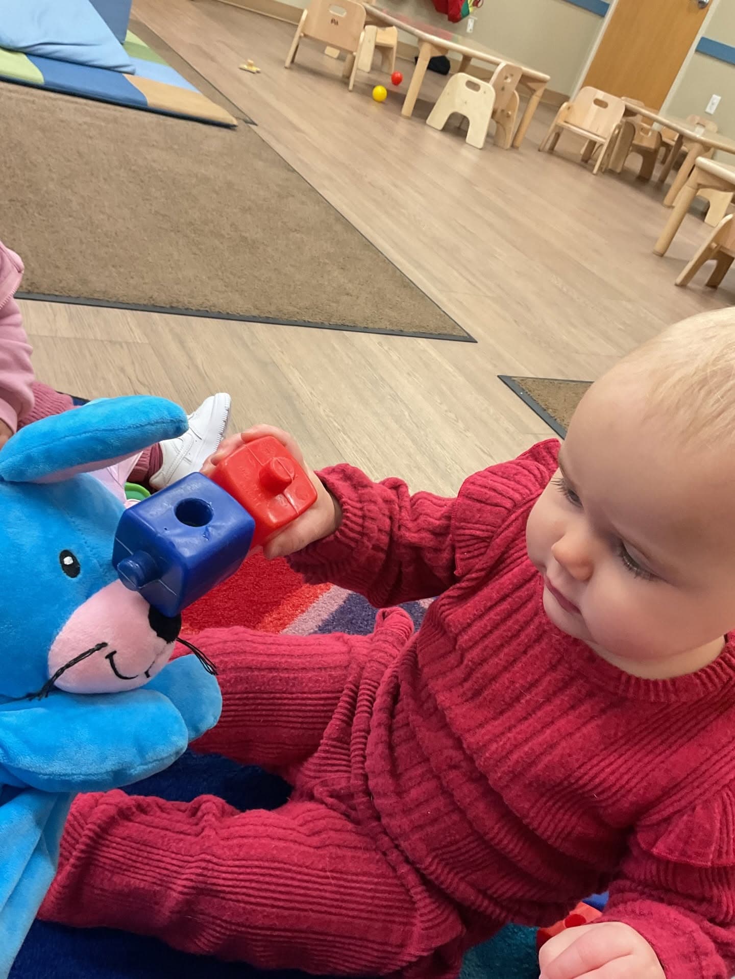 Infant in a red outfit stacking colorful blocks with Ally® the Bunny, developing hand-eye coordination and fine motor skills in the classroom.