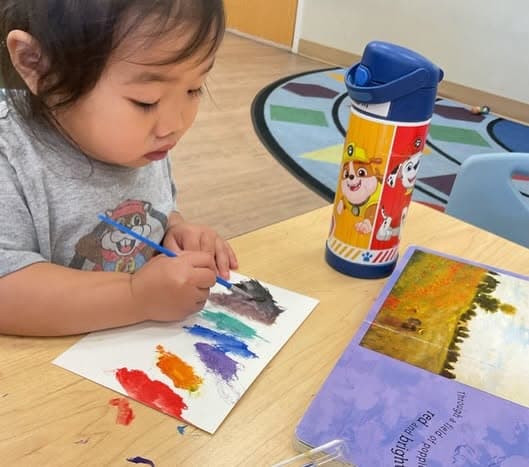 Young child painting a colorful rainbow of brushstrokes during a classroom art activity that encourages creativity, color recognition, and fine motor development.