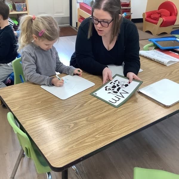 A teacher holding an "M" card and a child writing the letter "m"